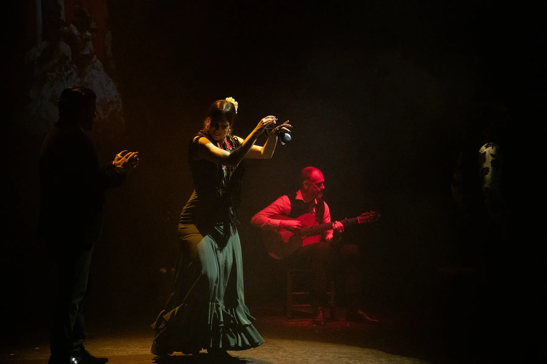 A dancer in a ruffled green dress raises castanets above her head while a guitarist in a red shirt plays behind her