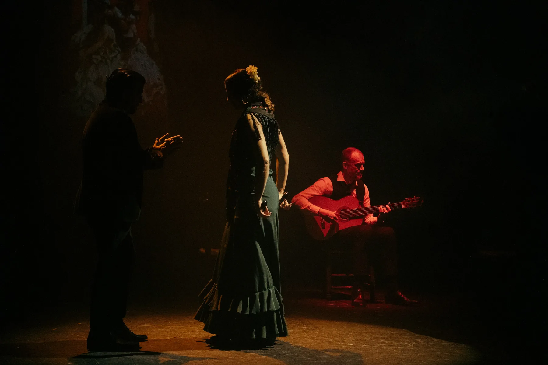 A dancer in a long green dress stands in a pool of warm light between a singer in silhouette and a guitarist in a red shirt
