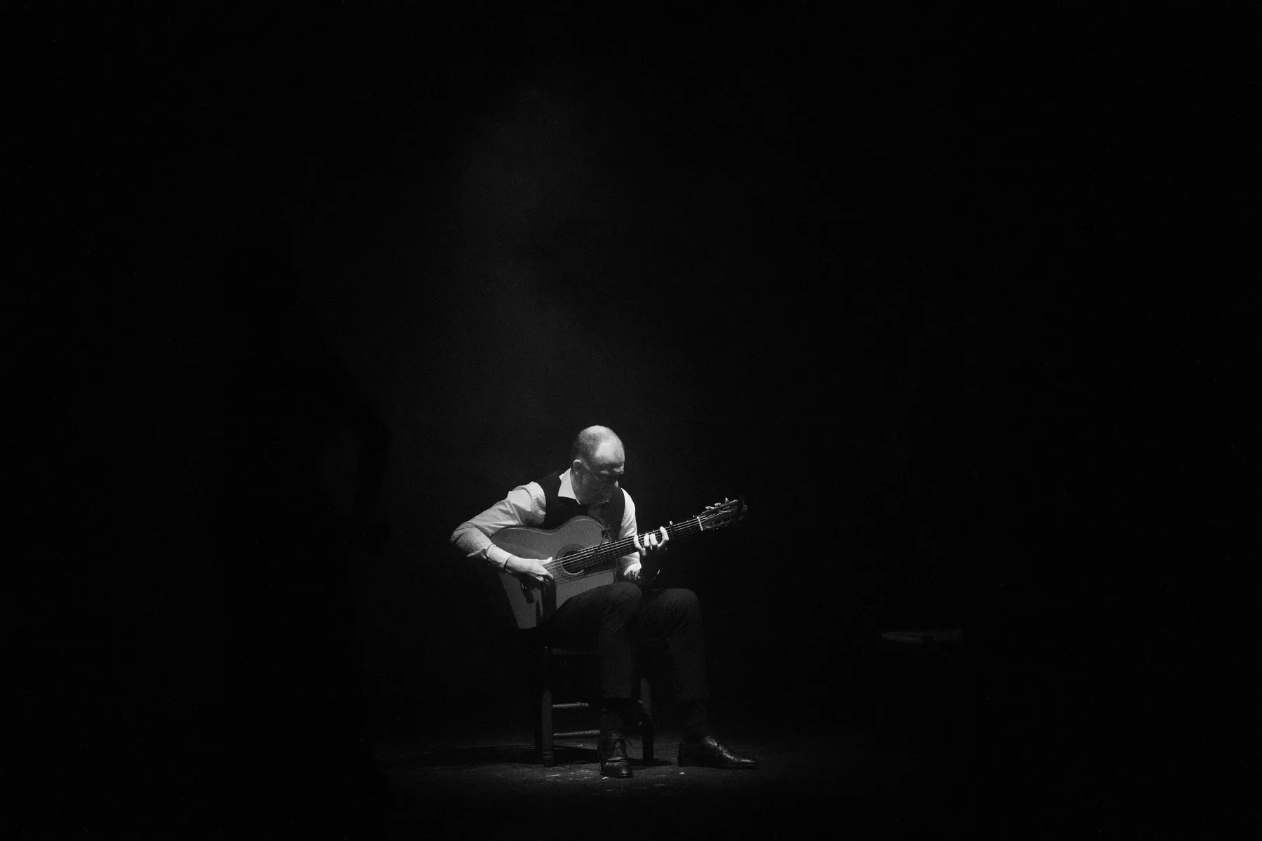 A guitarist sits alone in a pool of light on a dark stage, in black and white