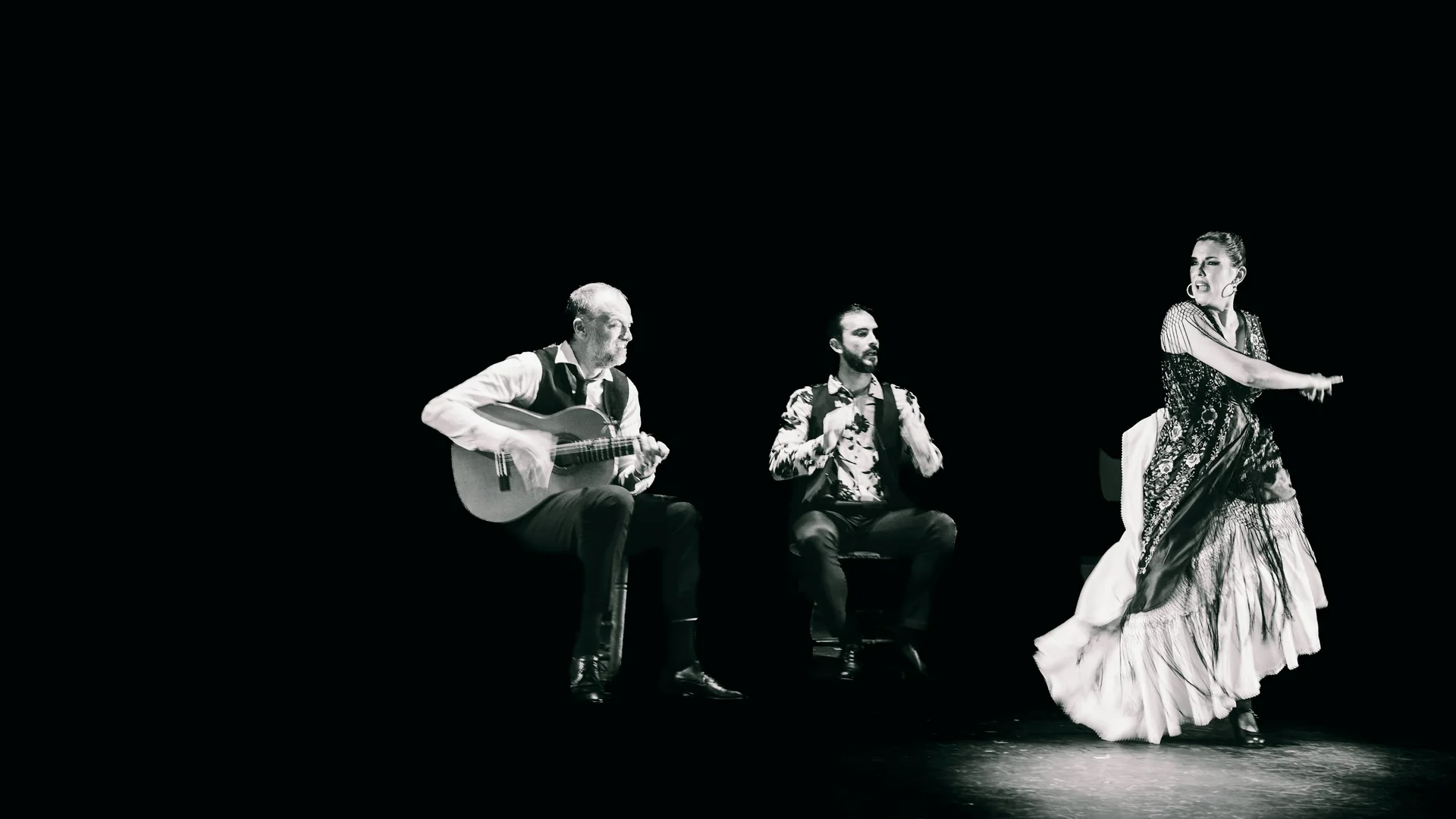 A flamenco dancer in a flounced white skirt and fringed shawl mid-turn beside a seated guitarist and a second musician, on a dark stage, in black and white